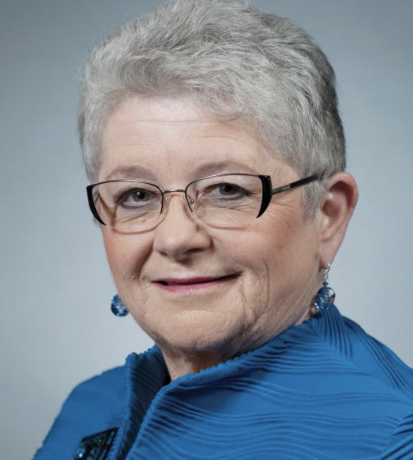 Professional headshot of Marcy Greenwood. She has short grey hair, glasses and is wearing a blue business coat and earings.